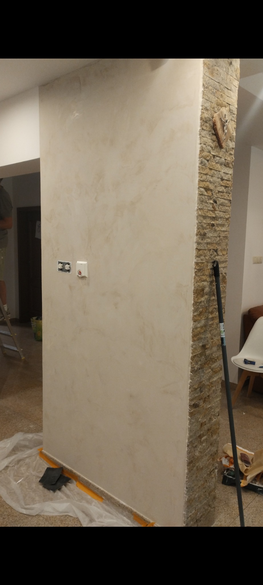 Ściana wewnętrzna po nałożeniu dekoracyjnej, beżowej farby strukturalnej, z widocznymi gniazdkami elektrycznymi i fragmentem kolumny wykończonej kamieniem dekoracyjnym. Na podłodze folia ochronna...