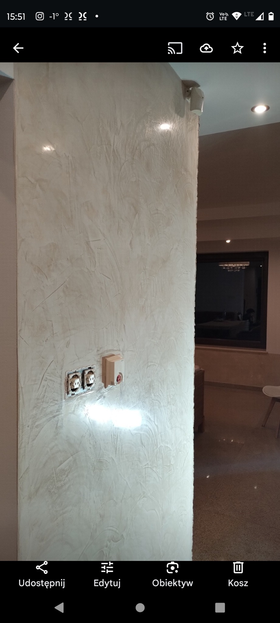 Ściana wewnętrzna wykończona dekoracyjnym tynkiem strukturalnym w odcieniach beżu i szarości, z widocznymi gniazdkami elektrycznymi i czujnikiem dymu, oświetlona przez wpadające światło odbijające...