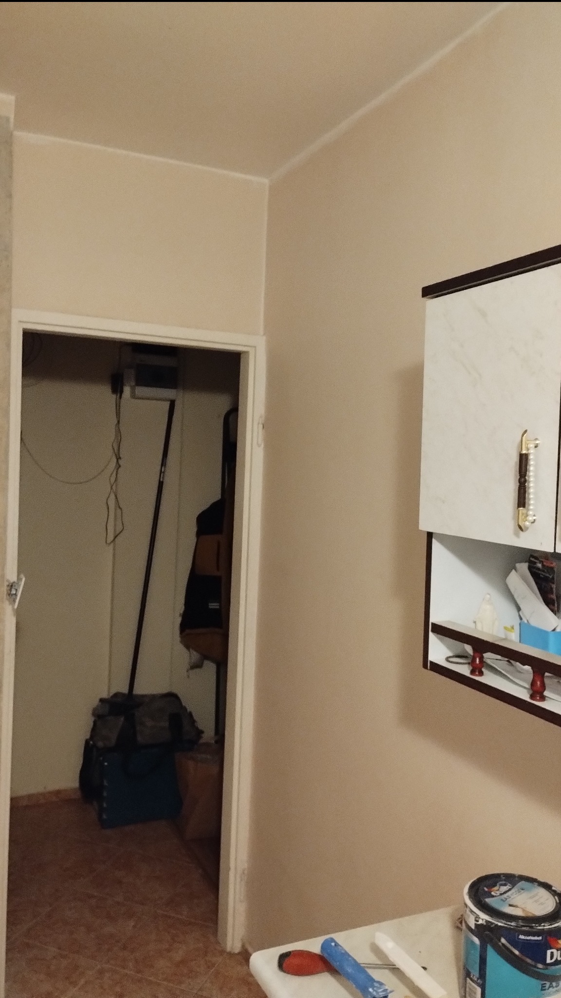 Ściana pomalowana na beżowo, widoczny fragment szafki wiszącej z ozdobnym uchwytem, otwarte drzwi do pomieszczenia z narzędziami i wiadro farby na blacie.