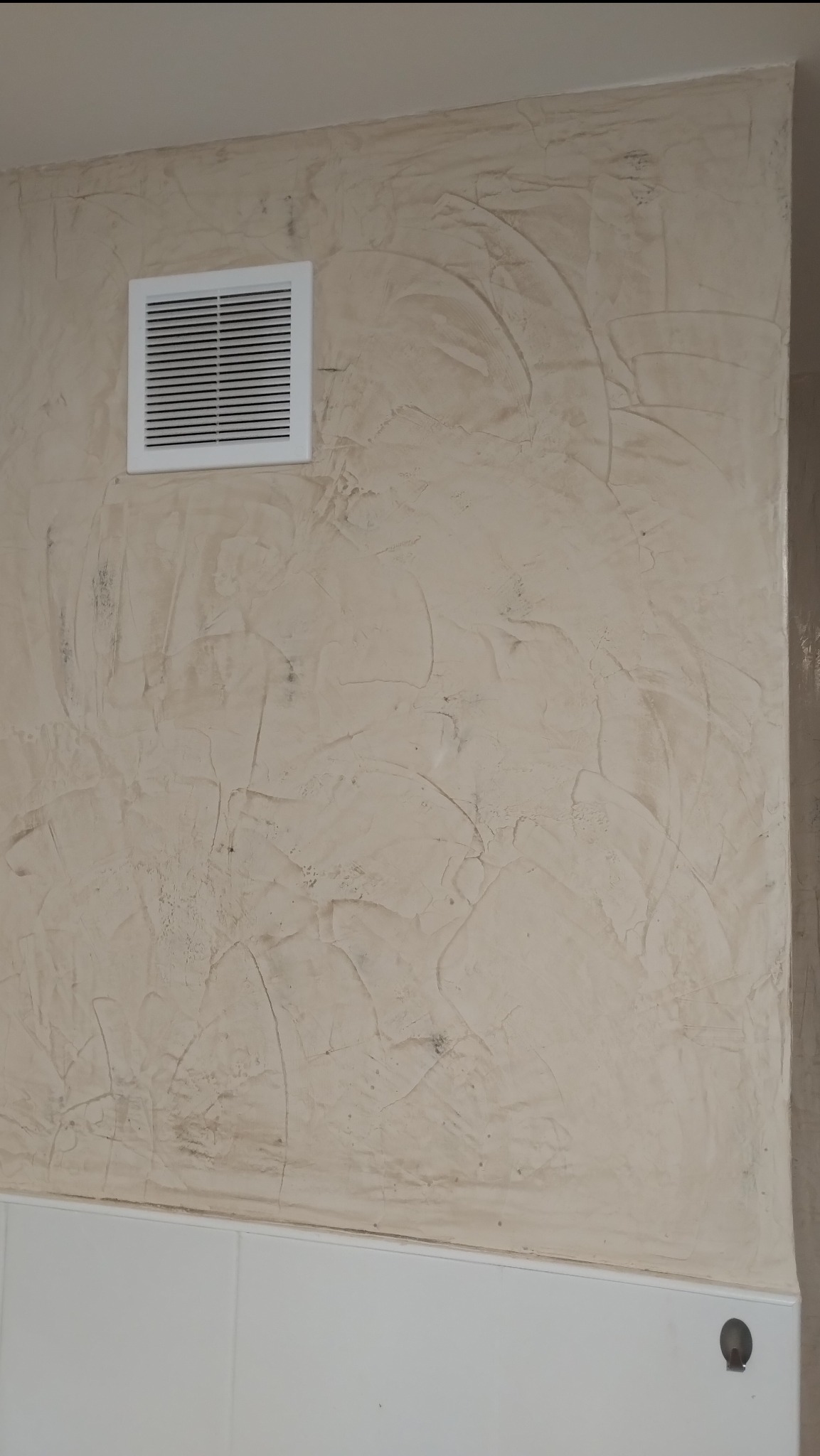 Ściana pokryta stiukiem weneckim w odcieniach beżu, z widoczną kratką wentylacyjną w górnej części i fragmentem białej lamperii na dole.