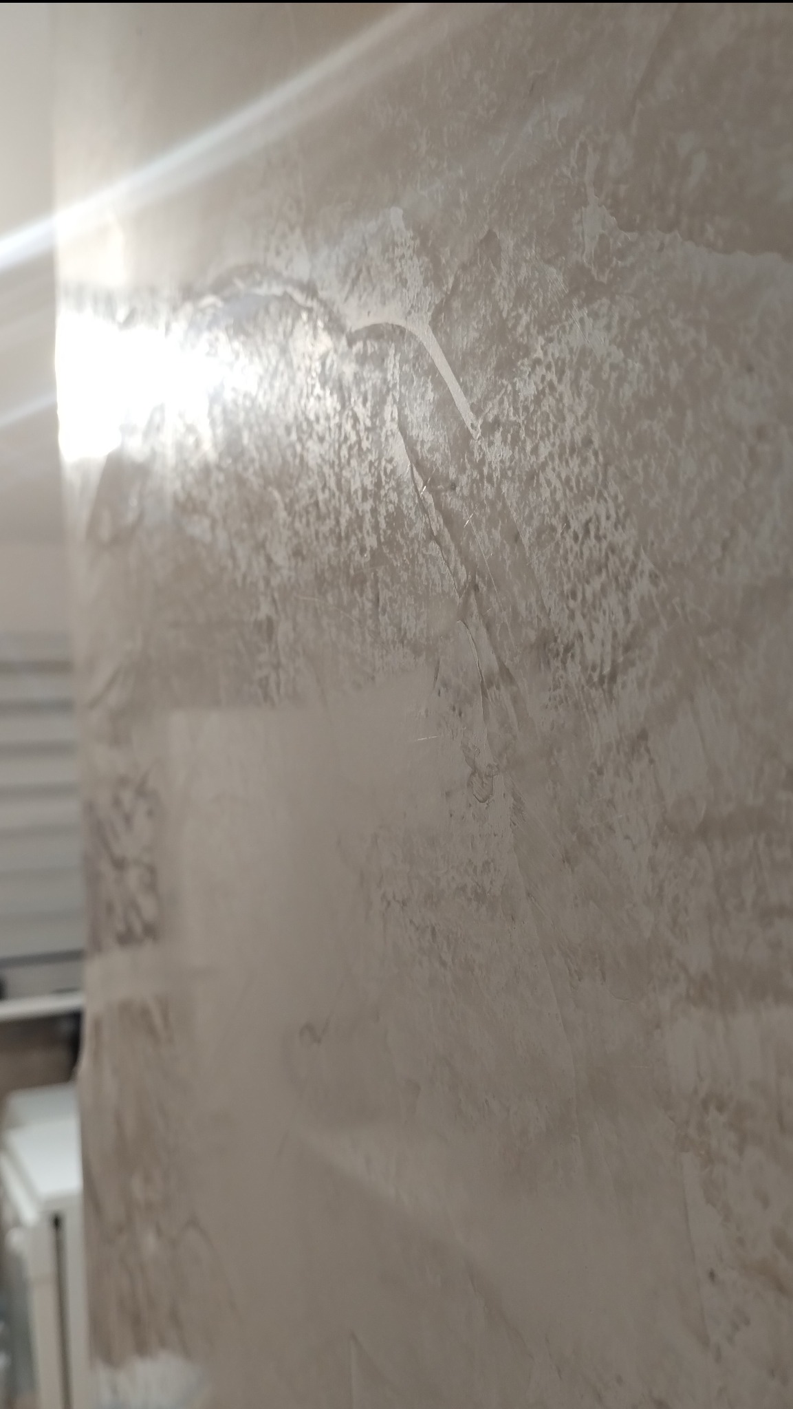 Ściana pokryta stiukiem weneckim Carmel, widoczne nieregularne wzory i faktura, delikatne refleksy światła, fragment białej szafki w tle.