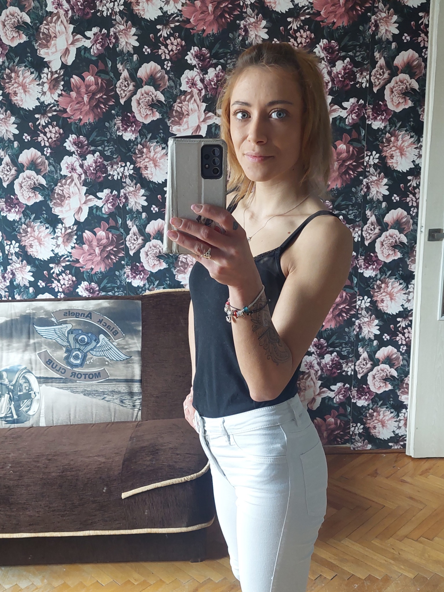 Kobieta robi selfie w mieszkaniu z ciemną tapetą w kwiaty i brązową kanapą z poduszką z motywem motocyklowym.