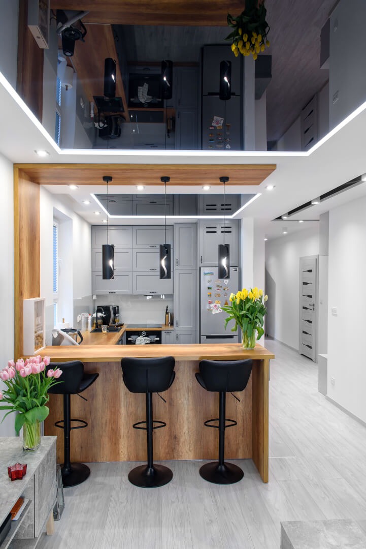 Nowoczesna kuchnia z wyspą i barowymi krzesłami, szare szafki, drewniany blat, czarne lampy wiszące, lustrzany sufit z oświetleniem LED, bukiet żółtych tulipanów i różowe tulipany w wazonie.