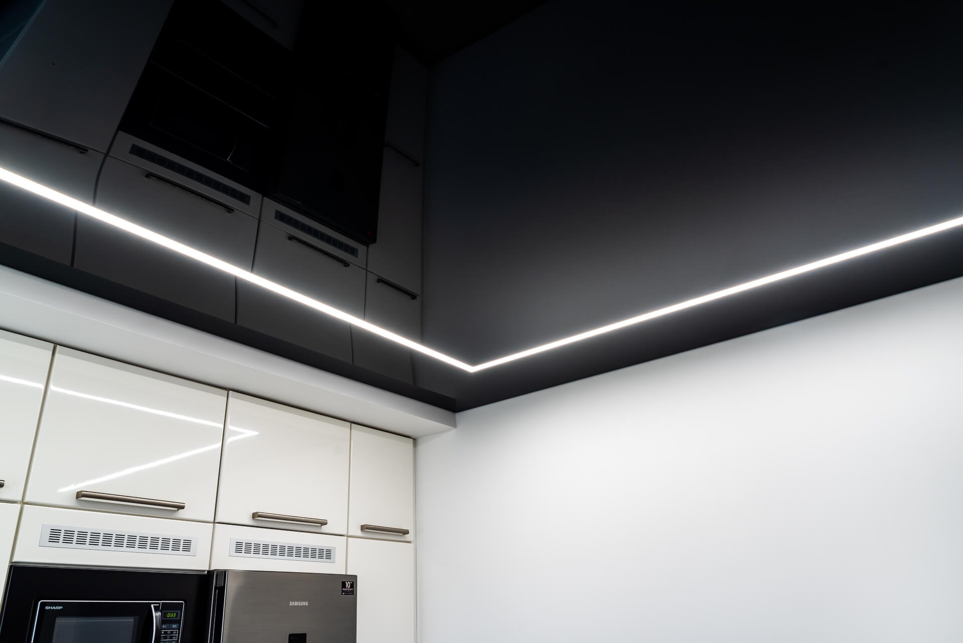 Czarne, lustrzane odbicie sufitu z liniowym oświetleniem LED w rogu, nad białą zabudową kuchenną.