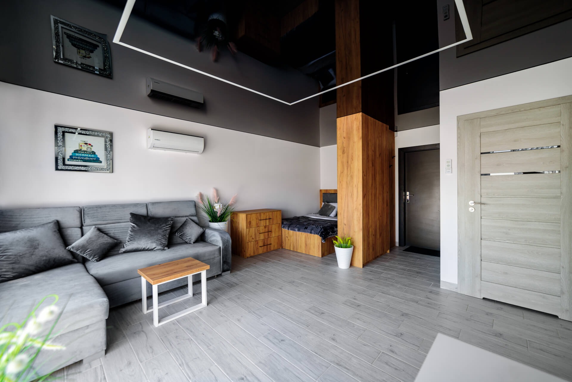 Nowoczesne wnętrze z szarą sofą, drewnianą zabudową dzielącą przestrzeń sypialną, błyszczącym czarnym sufitem i klimatyzacją, wykończone panelami podłogowymi imitującymi drewno.