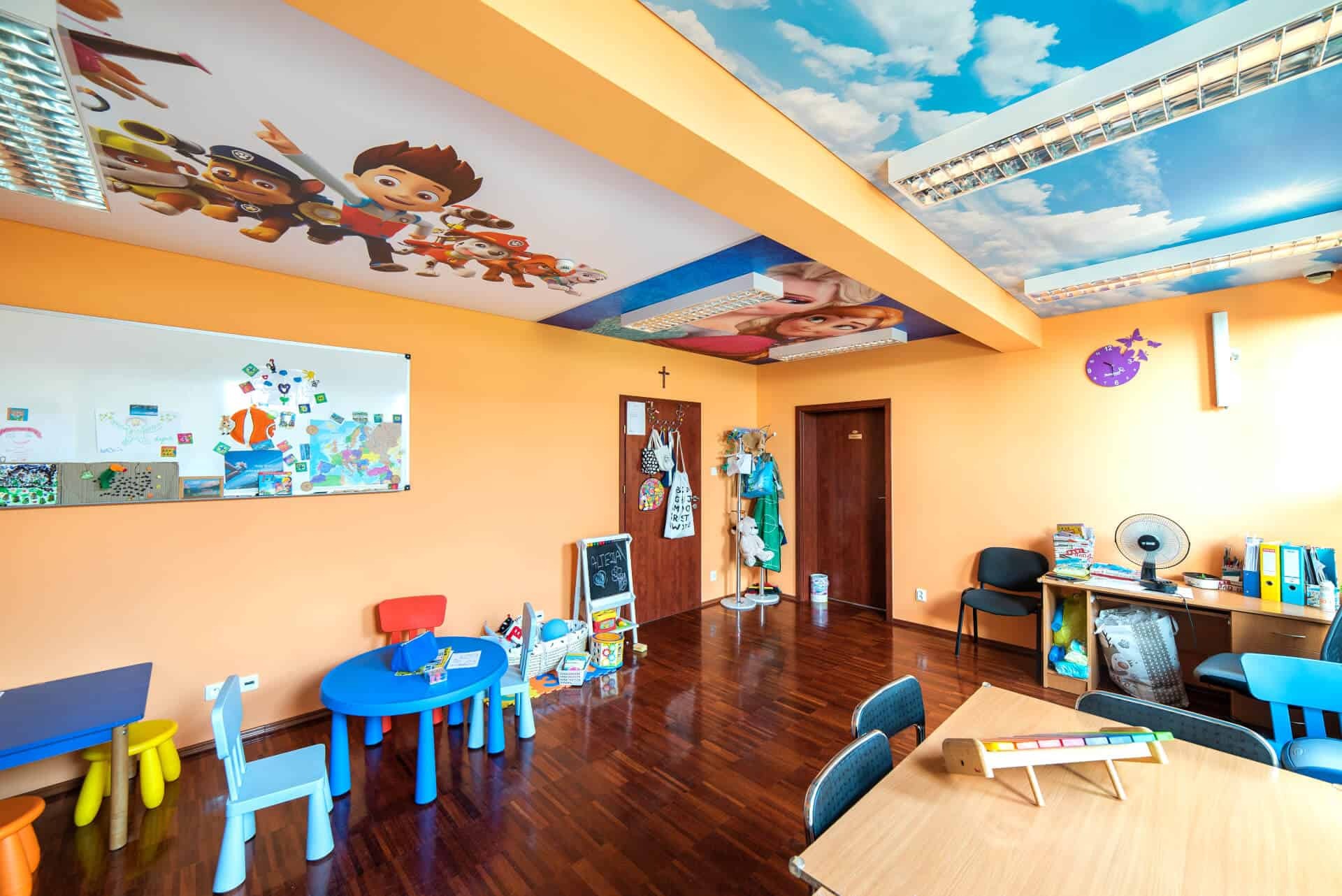 Wnętrze pokoju dziecięcego z pomarańczowymi ścianami, drewnianą podłogą i sufitem z motywem nieba i postaciami z bajek, wyposażone w kolorowe meble, białą tablicę i zabawki.