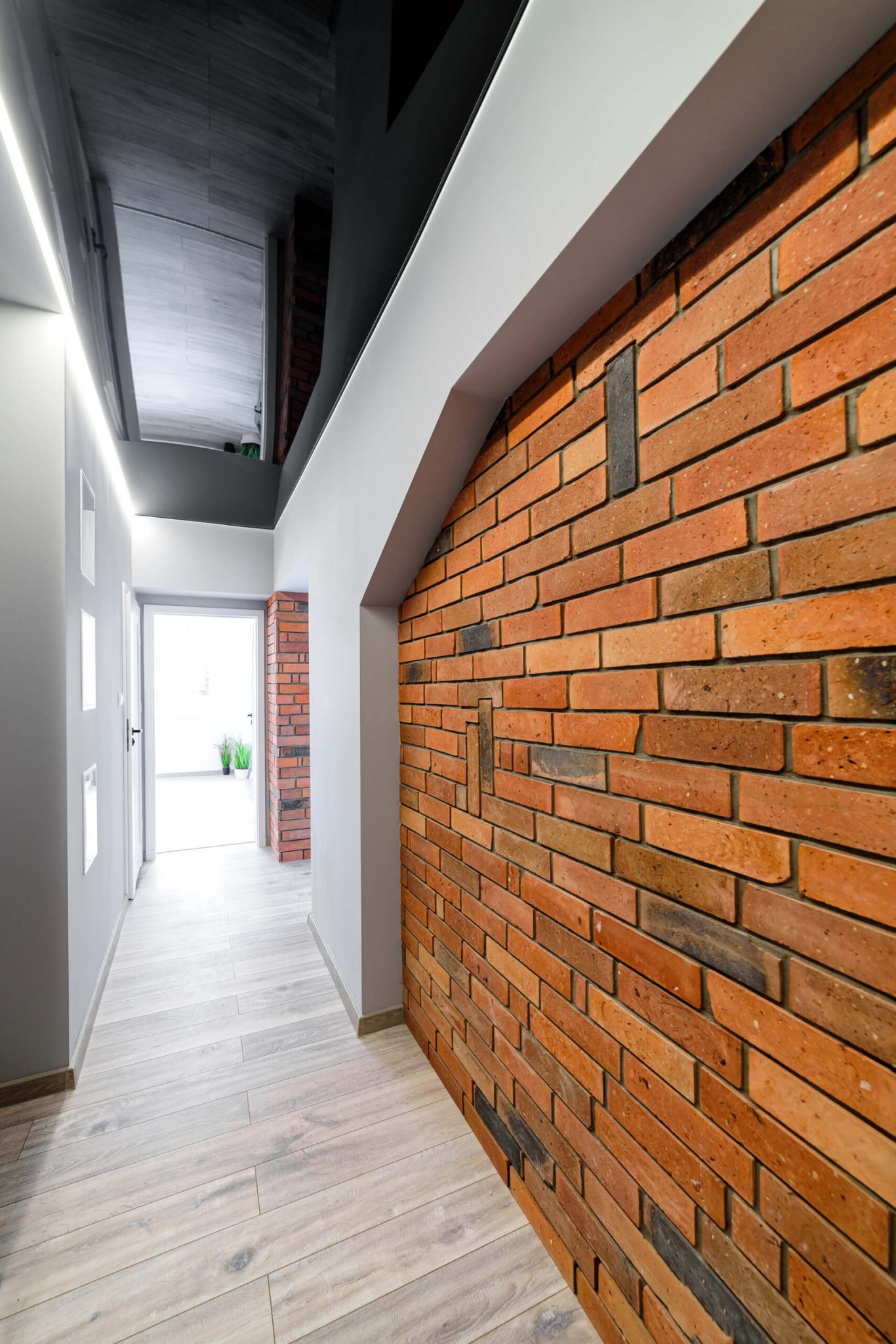 Korytarz z ceglaną ścianą w odcieniach czerwieni i brązu, drewnopodobna podłoga, białe ściany z wnękami i czarny, błyszczący sufit.