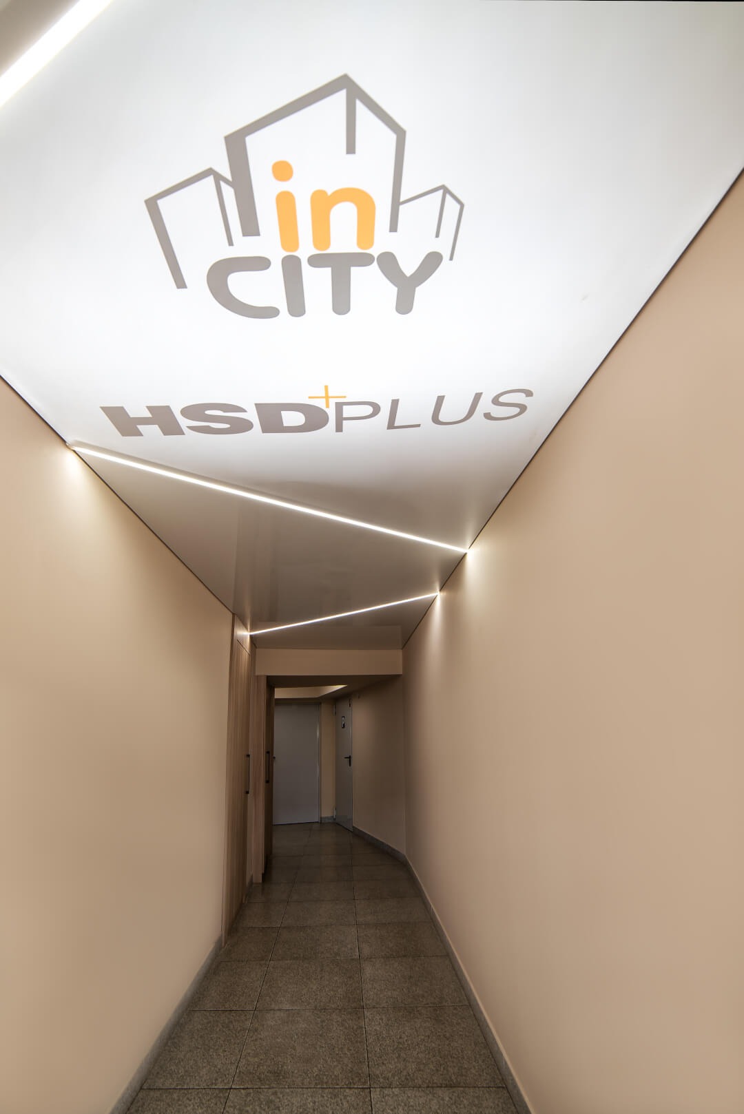 Korytarz biurowy z podwieszanym sufitem z logo firmy 'in CITY HSDplus' i liniowym oświetleniem LED, beżowe ściany i szara podłoga z płytek.