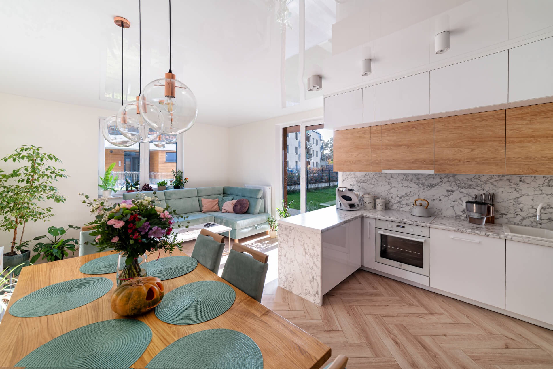 Jasne, przestronne wnętrze: kuchnia z białymi i drewnianymi szafkami, marmurowym blatem i podłogą w jodełkę, płynnie przechodząca w salon z turkusową sofą i drewnianym stołem z dekoracjami.