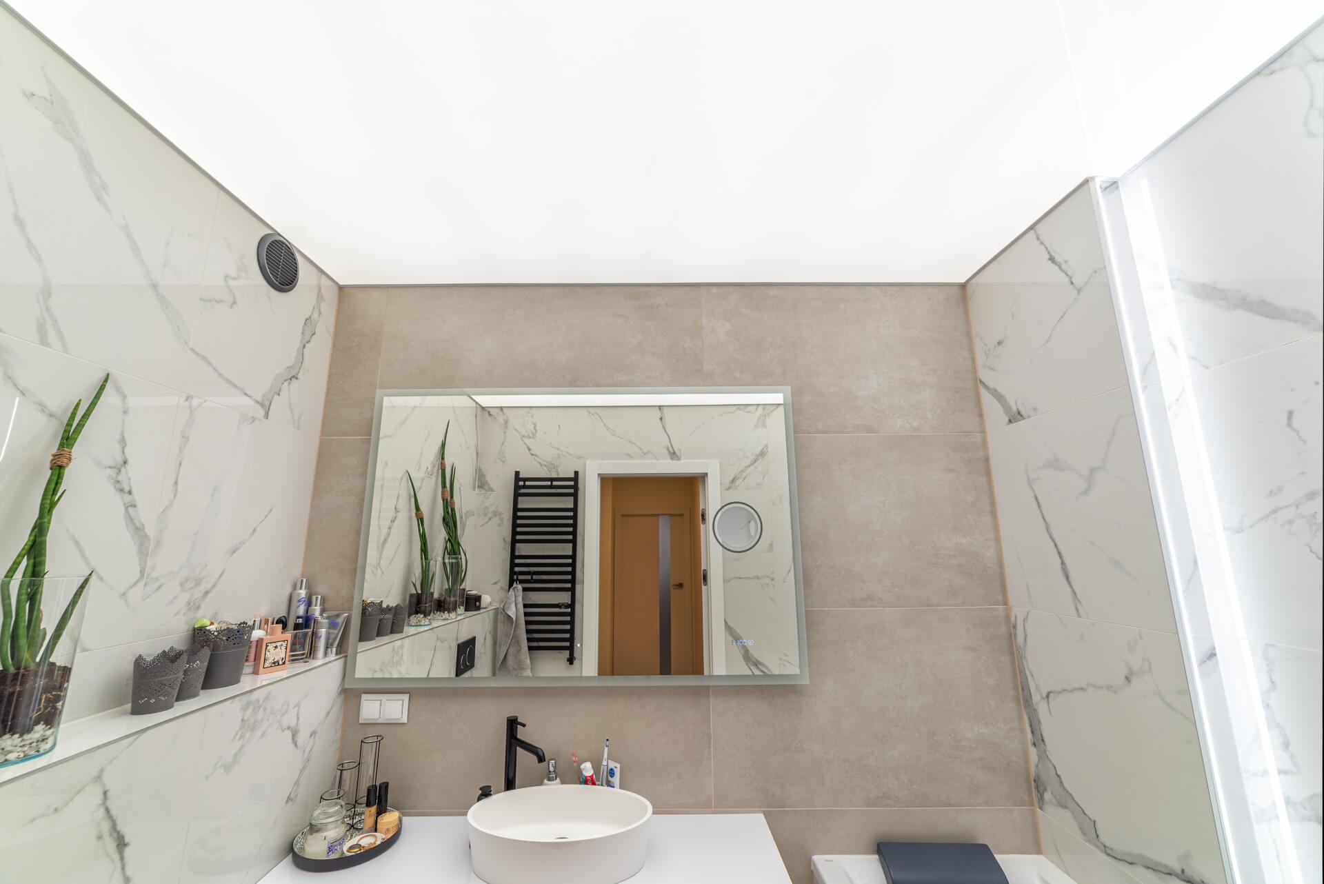 Nowoczesna łazienka z białymi marmurkowymi płytkami, beżowymi ścianami, podświetlanym sufitem, okrągłą umywalką nablatową i dużym lustrem z podświetleniem, w którym odbija się grzejnik drabinkowy...