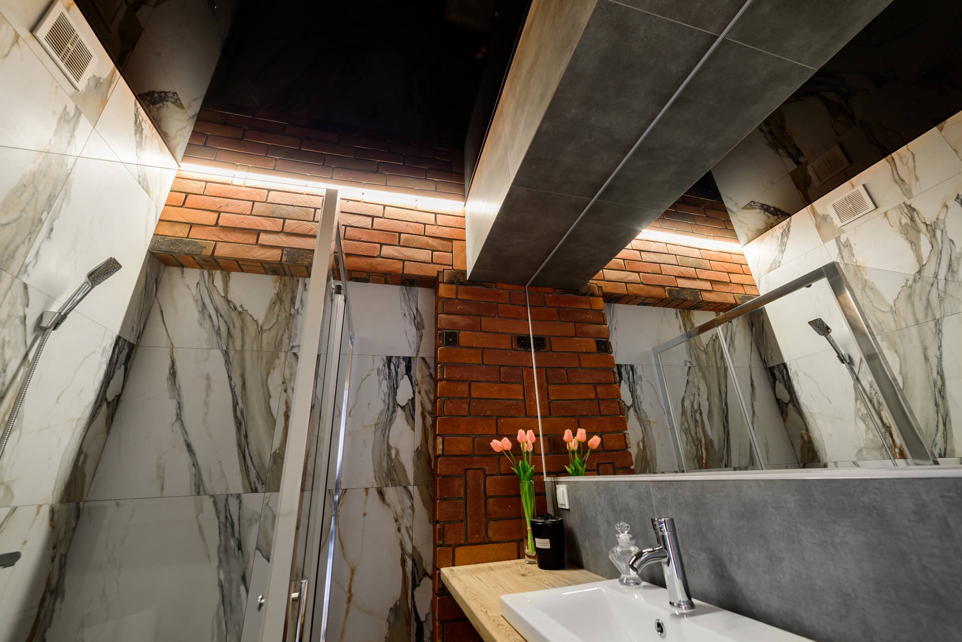Nowoczesna łazienka w Szczecinie z marmurowymi płytkami, ceglaną kolumną i podświetlonym sufitem. Widoczna kabina prysznicowa, umywalka z baterią i dekoracyjny wazon z tulipanami.