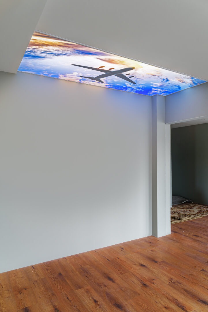 Sufit podwieszany z podświetleniem LED przedstawiający niebo z samolotem, drewniana podłoga, szare ściany, widok z perspektywy narożnika pokoju.