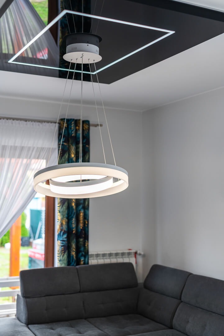 Nowoczesny okrągły żyrandol LED zawieszony pod czarnym, błyszczącym sufitem z kwadratowym oświetleniem LED, widoczny fragment szarej sofy i okna z wzorzystymi zasłonami.