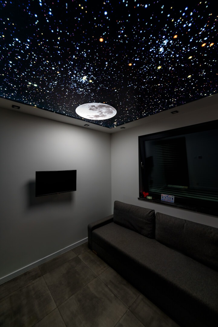 Sufit napinany z motywem kosmosu i podświetlaną imitacją księżyca w pokoju z szarą sofą i telewizorem na ścianie.