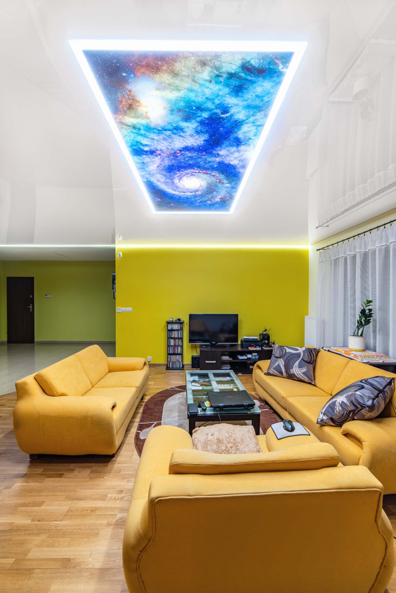 Salon z żółtymi ścianami i sofami, podświetlany sufit z motywem kosmosu, drewniana podłoga.