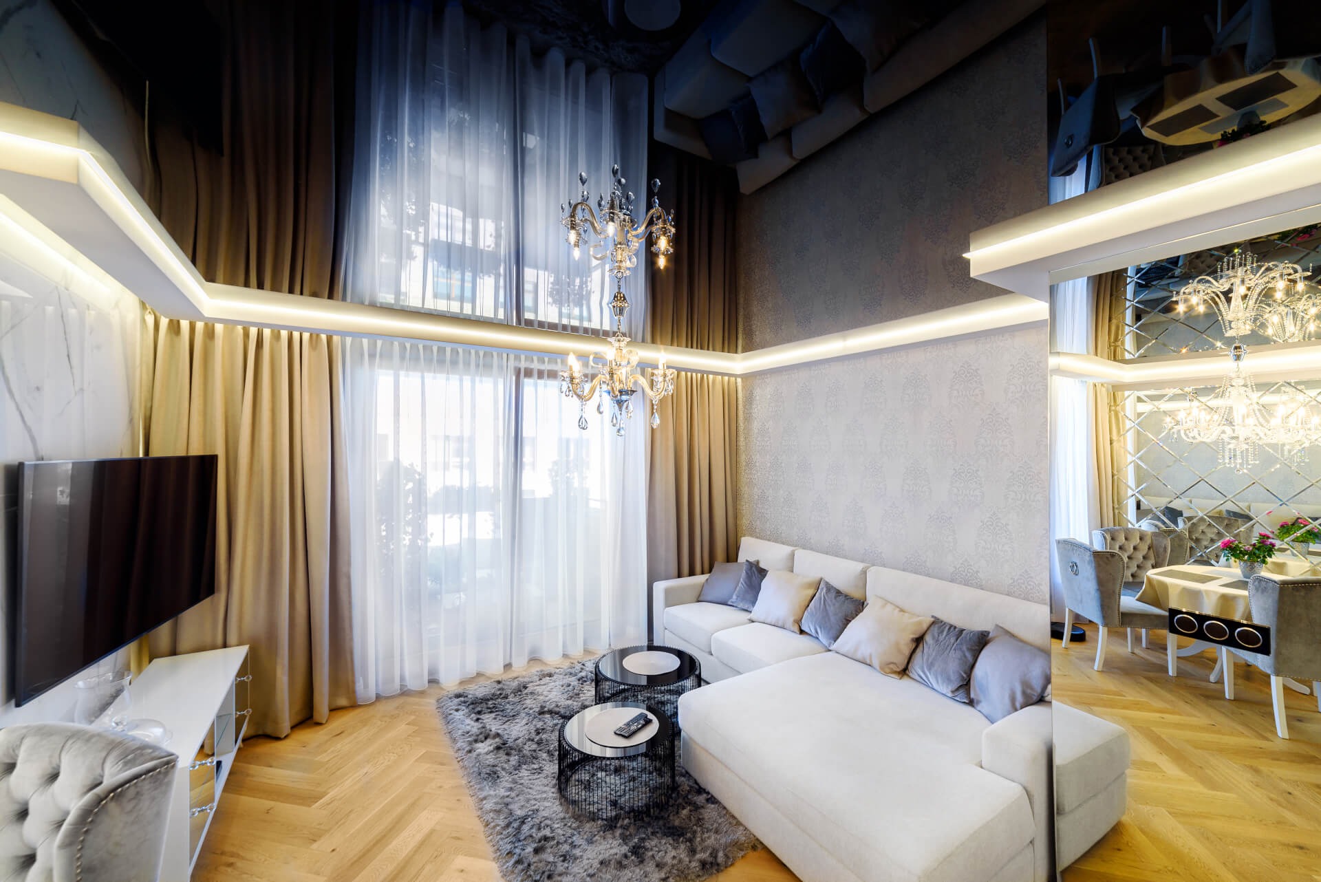 Elegancki salon z jasną sofą narożną, okrągłymi stolikami kawowymi na dywanie shaggy i kryształowym żyrandolem, oświetlenie LED w suficie i marmurowa ściana z telewizorem.