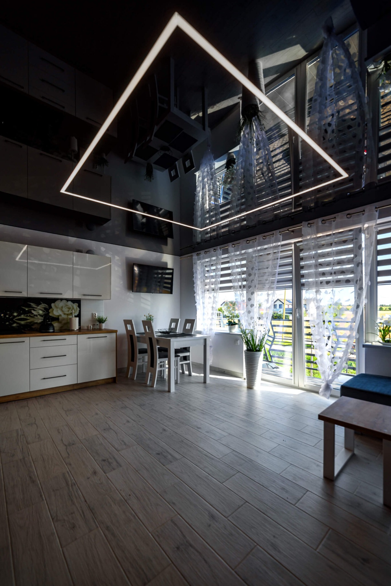 Nowoczesne wnętrze z czarnym, lustrzanym sufitem i kwadratową lampą LED, biała kuchnia z drewnianym blatem, stół z krzesłami, okno z roletami i firankami.