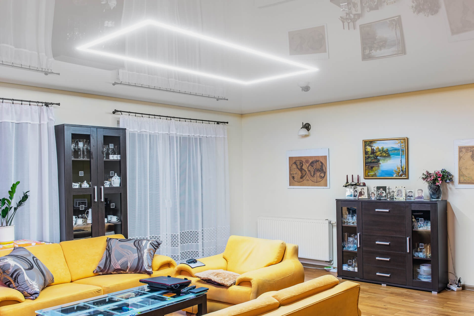 Salon z żółtymi sofami i podświetlanym, kwadratowym sufitem napinanym z połyskiem, odbijającym elementy wyposażenia.