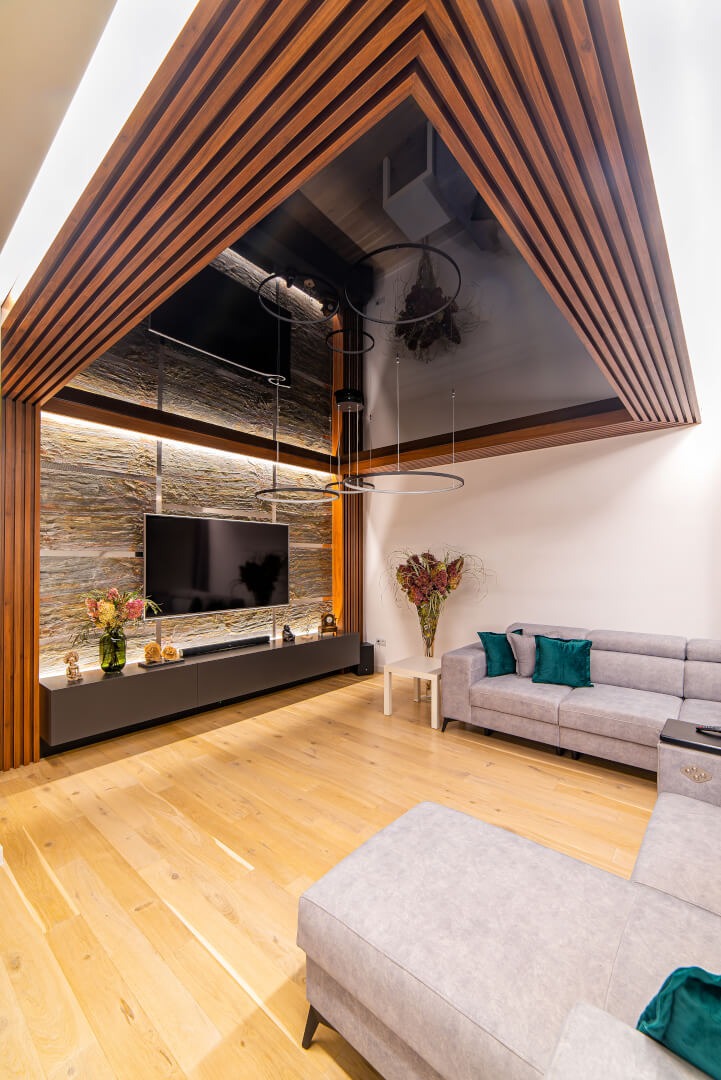 Salon z drewnianym sufitem kasetonowym, czarnym sufitem napinanym z odbiciami, kamienną ścianą akcentującą podświetloną LED, szarą sofą narożną i drewnianą podłogą.