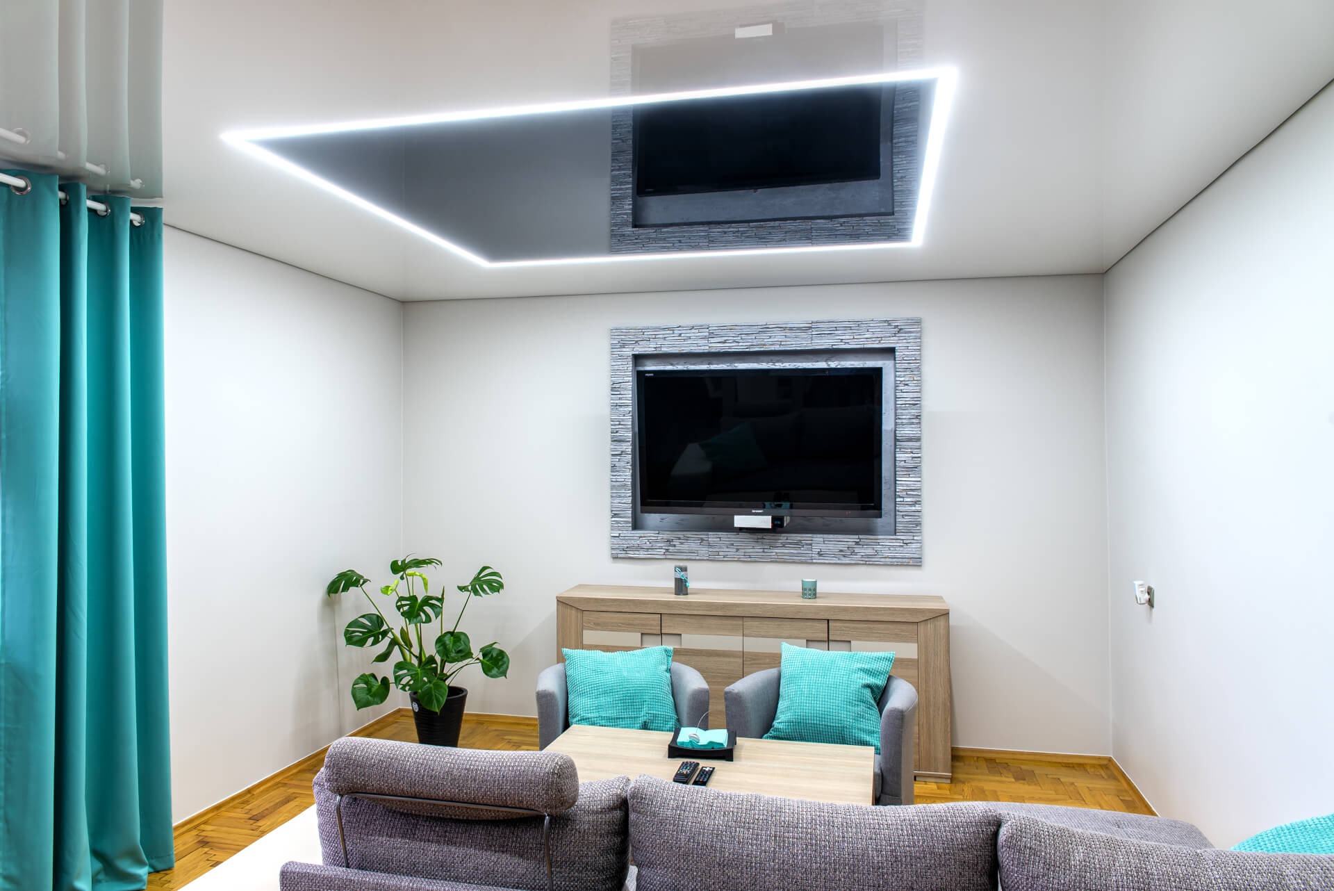 Nowoczesny salon z podwieszanym sufitem z czarną wstawką i oświetleniem LED, telewizor w ramie z imitacji kamienia, turkusowe zasłony i poduszki, szara sofa i drewniana podłoga.