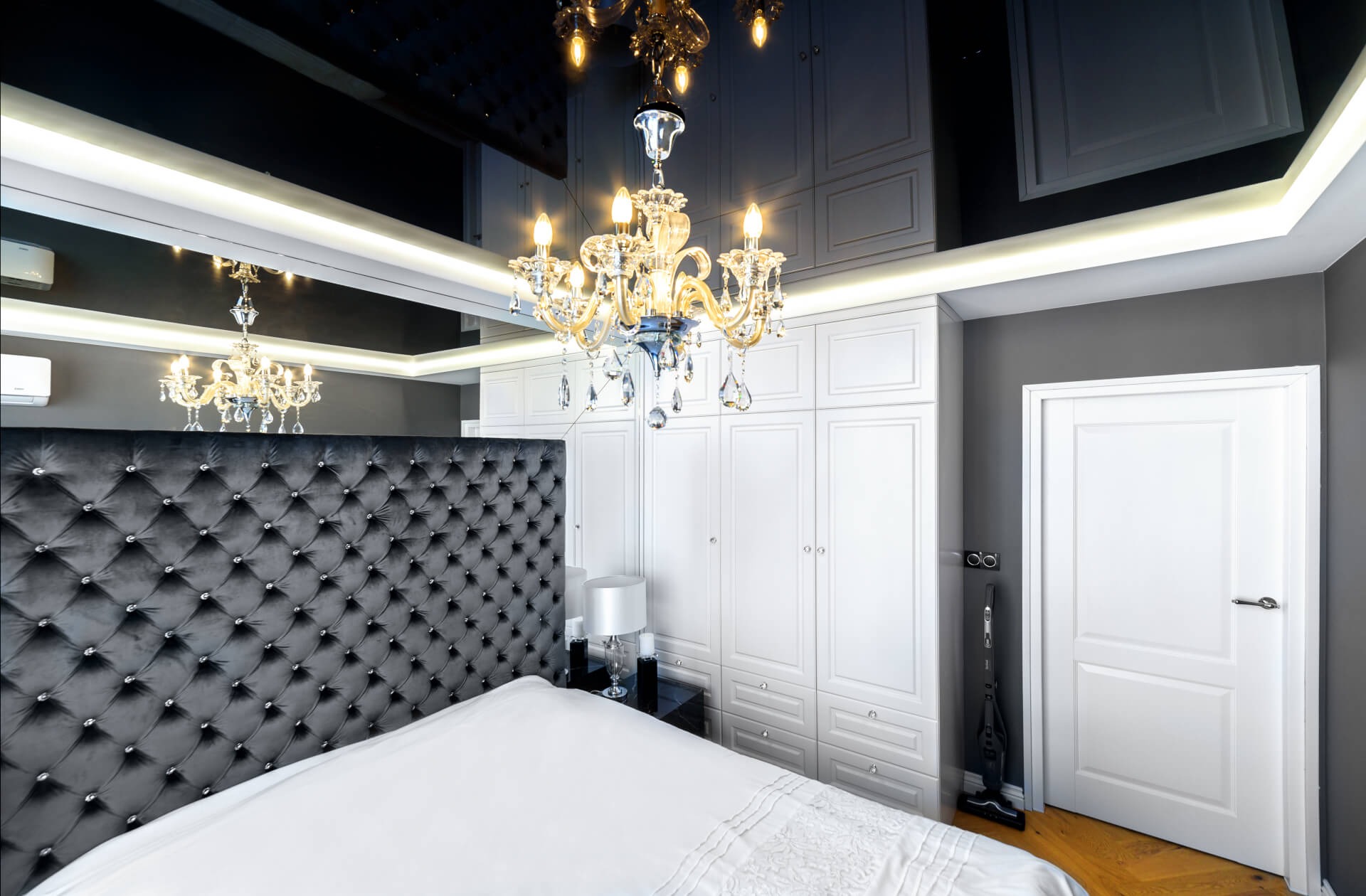Sypialnia z czarnym, pikowanym zagłówkiem, białymi szafami w zabudowie, błyszczącym czarnym sufitem z oświetleniem LED i kryształowym żyrandolem.