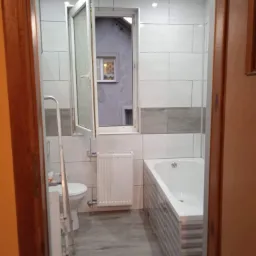 Wykończona łazienka z biało-szarymi płytkami na ścianach i podłodze, widoczna wanna, toaleta, drabina malarska i okno z widokiem na szary budynek.