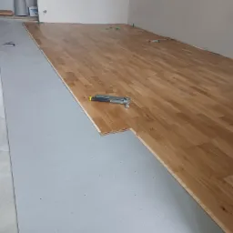 Układanie drewnianej podłogi: widoczne deski parkietu, podkład izolacyjny w kolorze szarym, narzędzie do montażu i zielone kliny dystansowe przy ścianie.