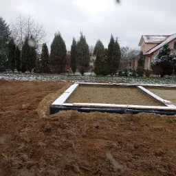 Przygotowany teren pod fundamenty z widoczną izolacją przeciwwilgociową i drewnianym szalunkiem, częściowo pokryty śniegiem, na tle domu jednorodzinnego i drzew w zimowej scenerii.