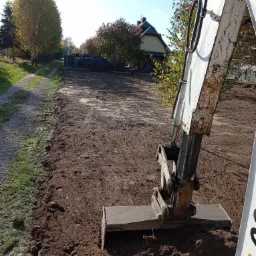Biała minikoparka z łyżką wyrównuje teren działki w słoneczny dzień, widoczny świeżo uprawiony grunt, drzewa w tle i częściowo zabudowania.