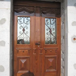 Dwuskrzydłowe, ciemnobrązowe drzwi wejściowe z przeszkleniami ozdobionymi metalowymi, kręconymi elementami, zamontowane w surowym otworze ściany z betonu komórkowego.