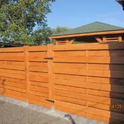 Nowoczesne, poziome ogrodzenie z drewna w ciepłym odcieniu brązu, z widocznymi zawiasami i fragmentem furtki, zamontowane na betonowej podmurówce, z fragmentem altany w tle.