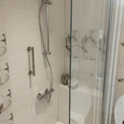 Wykończona kabina prysznicowa z chromowaną armaturą, szklaną ścianką i beżowymi płytkami z motywem kwiatowym, uzupełniona o dekoracyjny słoik z muszelkami i dozownik mydła.