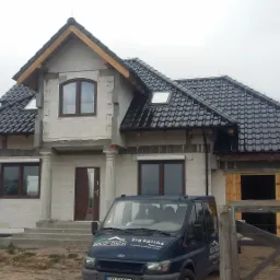 Dom jednorodzinny w stanie surowym z czarną dachówką ceramiczną, widoczne rynny, okna dachowe i samochód dostawczy z logo firmy dekarskiej zaparkowany przed budynkiem.