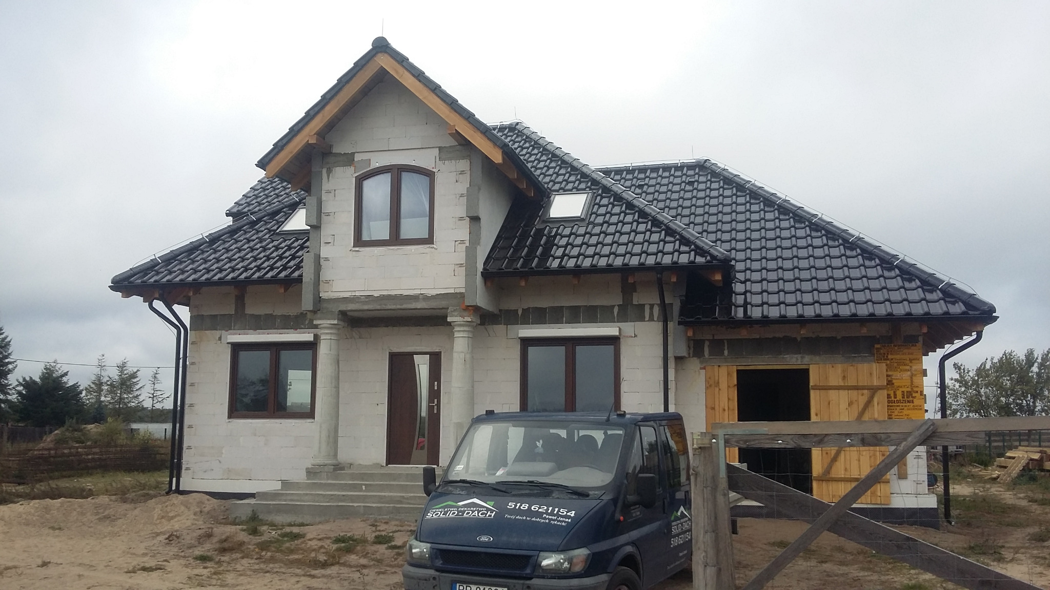 Dom jednorodzinny w stanie surowym z czarną dachówką ceramiczną, widoczne rynny, okna dachowe i samochód dostawczy z logo firmy dekarskiej zaparkowany przed budynkiem.