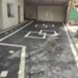 Ułożona kostka brukowa w kolorze grafitowym z jasnymi, kwadratowymi wstawkami tworzącymi dekoracyjny wzór na podjeździe przed budynkiem z zadaszeniem.