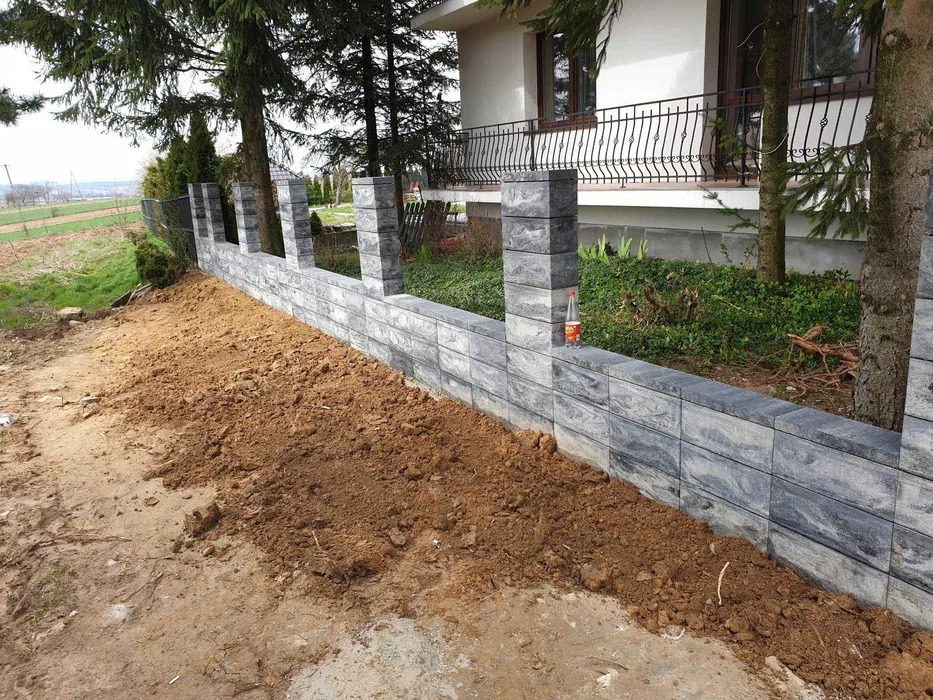Niski murek ogrodzeniowy z szarej kostki betonowej z filarami, świeżo obsypany ziemią, z butelką wody mineralnej na górze, na tle domu z balkonem.