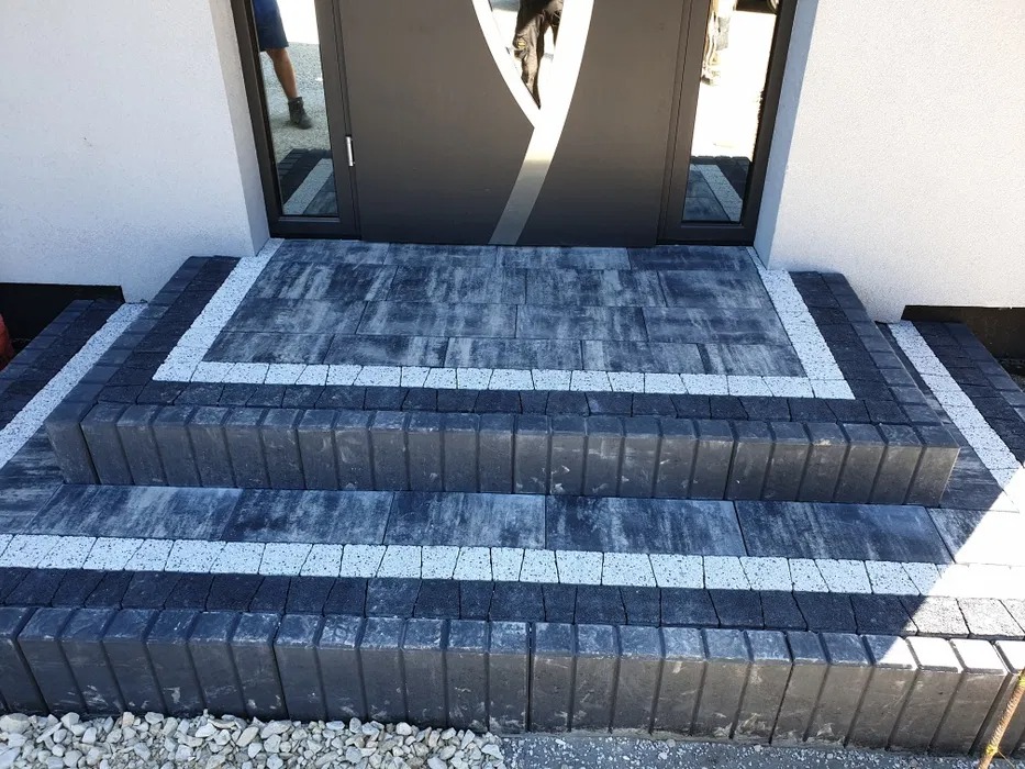 Schody wejściowe wyłożone ciemną kostką brukową z jasnymi akcentami, prowadzące do nowoczesnych drzwi wejściowych z elementami szkła. Widoczne detale wykończenia i małe kamyczki u podstawy schodów.