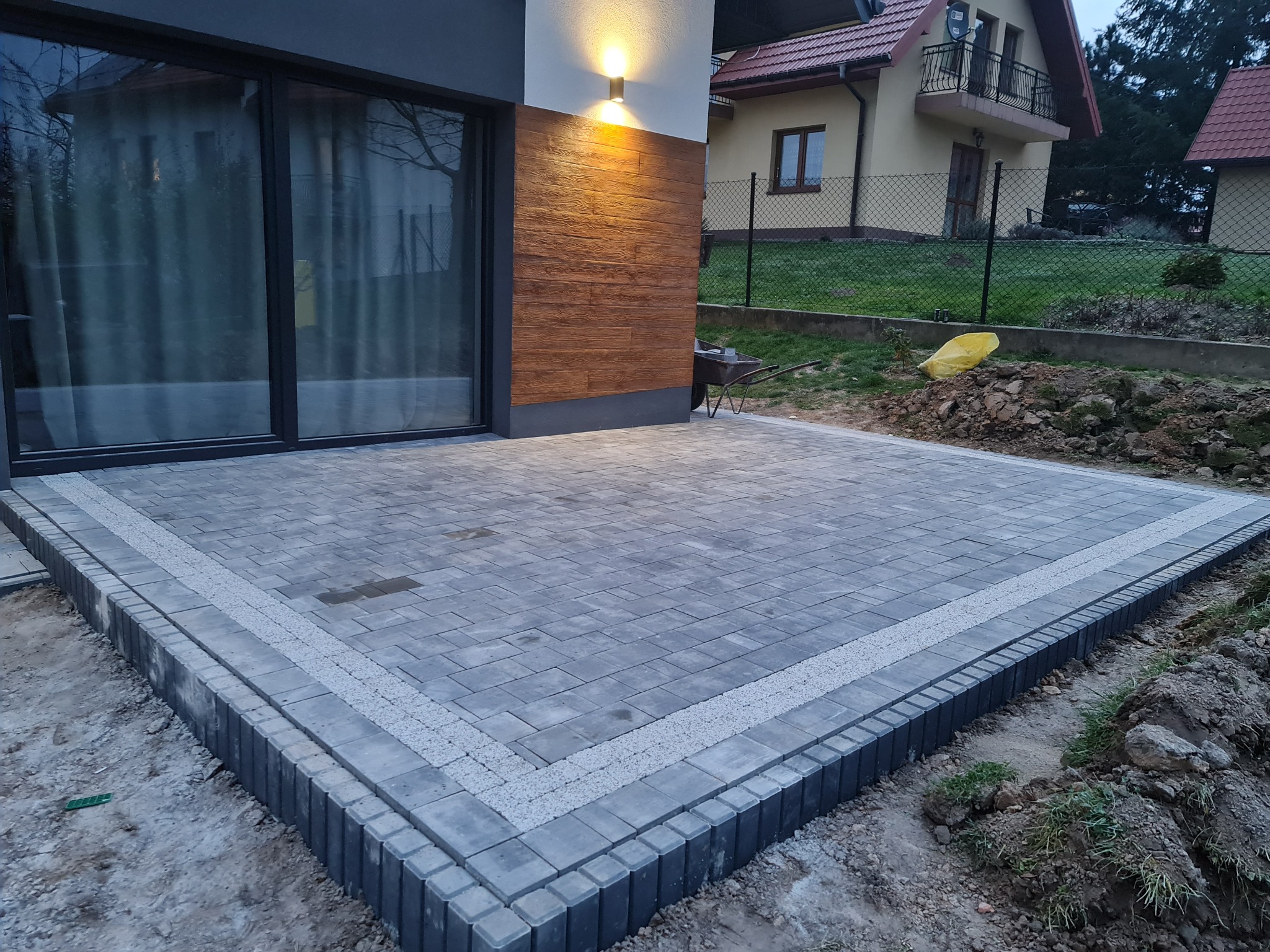 Nowoczesny taras z szarej kostki brukowej ułożonej na planie kwadratu, obrzeża z ciemniejszych kostek z wypełnieniem z jasnego grysu, widok na dom z dużymi oknami i elewacją z imitacji drewna.