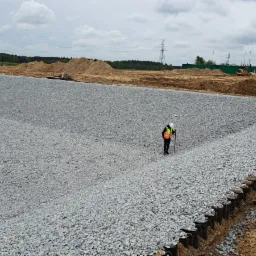 Pomiar terenu pod zbiornik retencyjny z szarego kruszywa, pracownik w kamizelce odblaskowej używa sprzętu pomiarowego na tle hałd piasku i drzew.