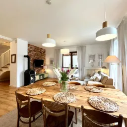 Jasne wnętrze domu z drewnianym stołem, krzesłami, kominkiem z ceglaną ścianą i schodami w tle. Na stole dekoracyjne podkładki i wazon z kwiatami.