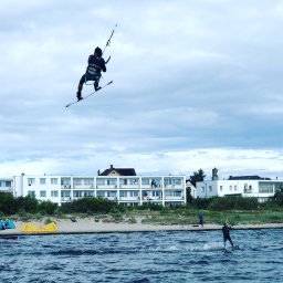Michal_Cedro - Kitesurfer w locie nad wodą w Warszawie, w tle budynek mieszkalny i plaża.