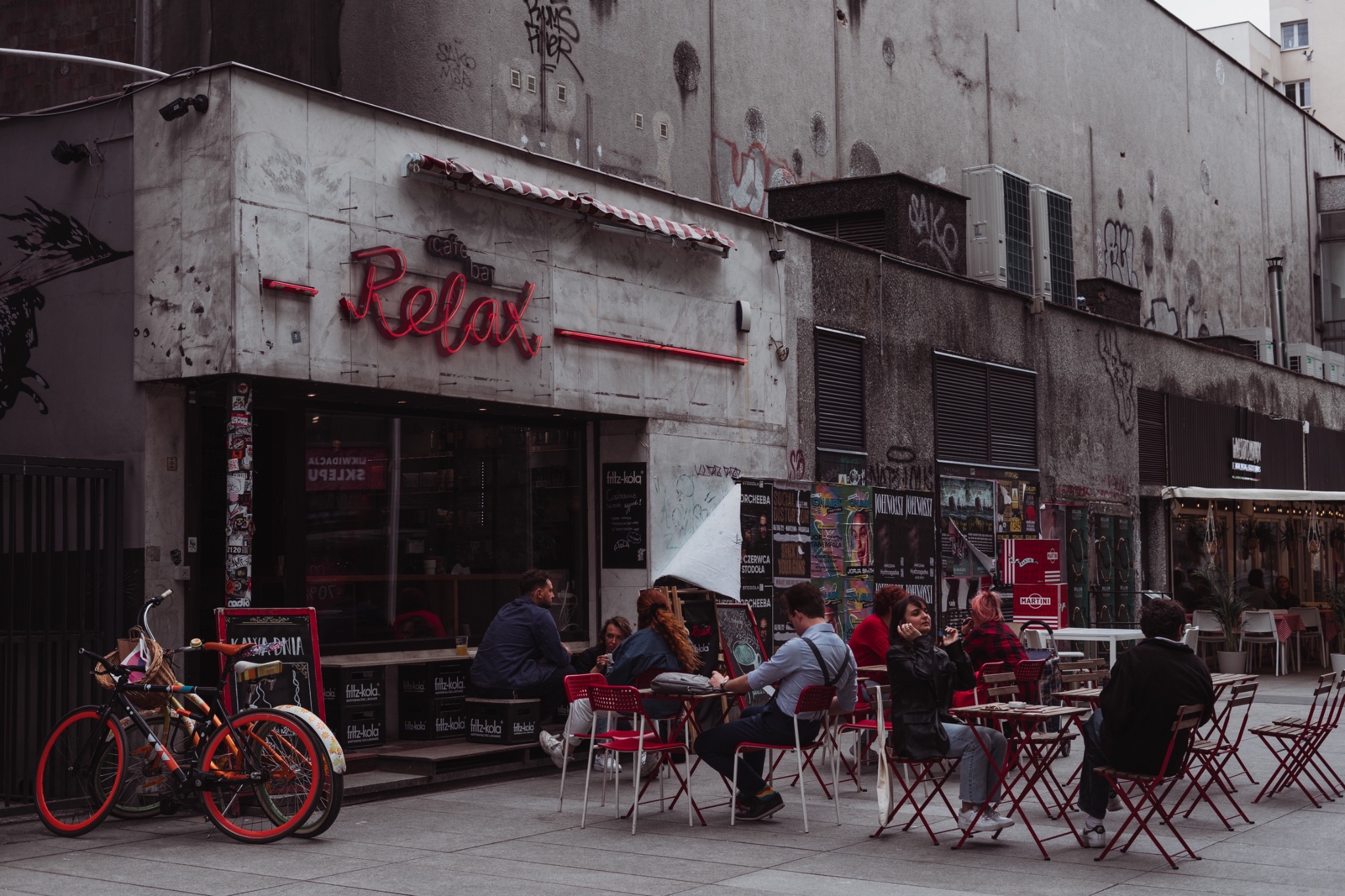 Kawiarnia z neonowym napisem 'Relax' i stolikami na zewnątrz, gdzie siedzą ludzie. Dwa rowery zaparkowane obok wejścia, ściany budynku pokryte graffiti i plakatami.