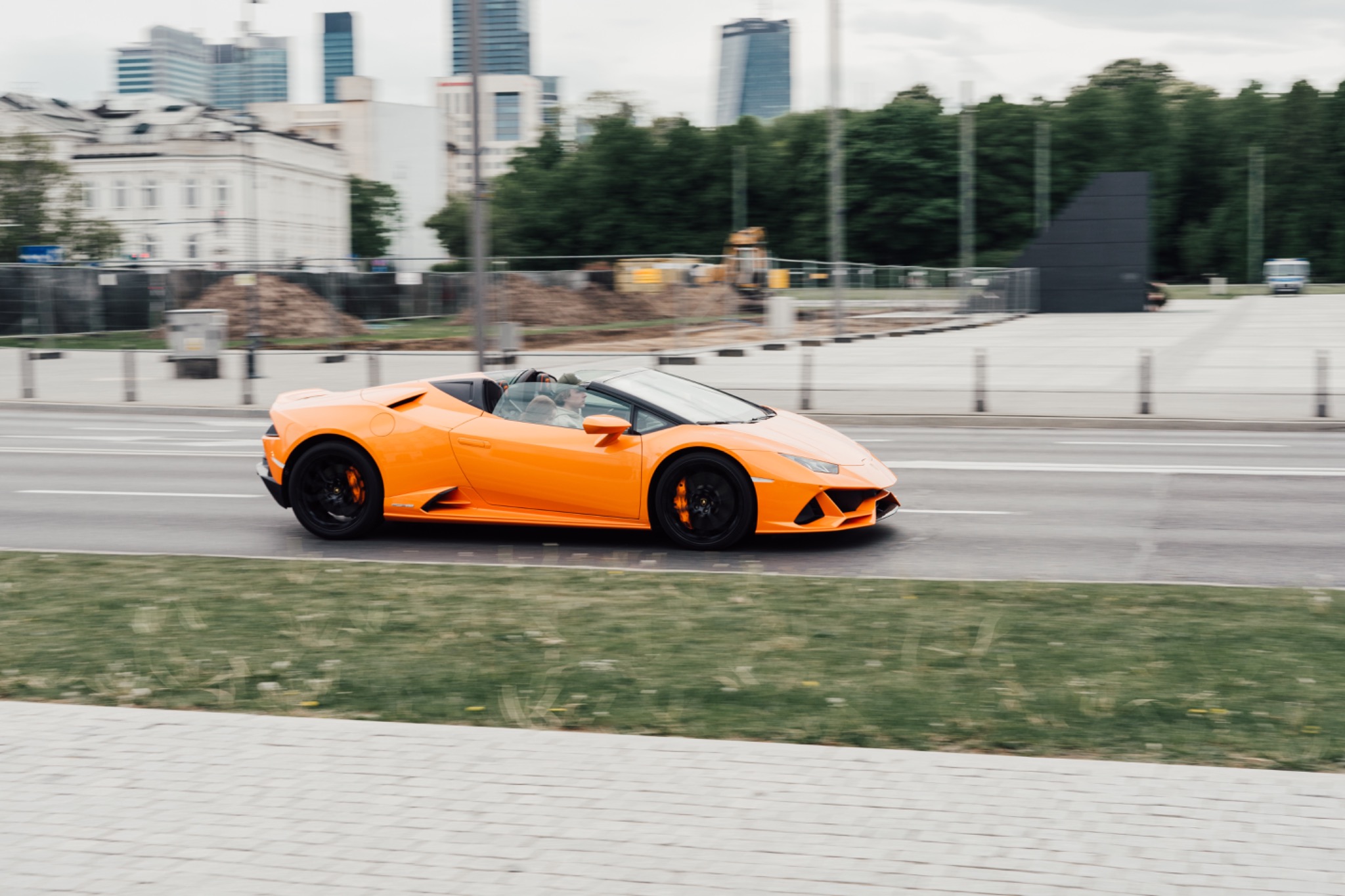 Pomarańczowy Lamborghini Huracan Spyder pędzi ulicą na tle budynków Warszawy, z widocznymi czarnymi felgami i zaciskami hamulcowymi w kolorze nadwozia.
