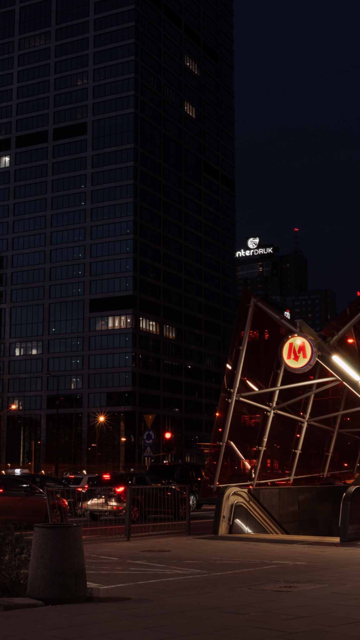 Nocne ujęcie wejścia do metra z charakterystycznym czerwonym logo 'M' na tle nowoczesnego budynku biurowego z widocznymi światłami w oknach.