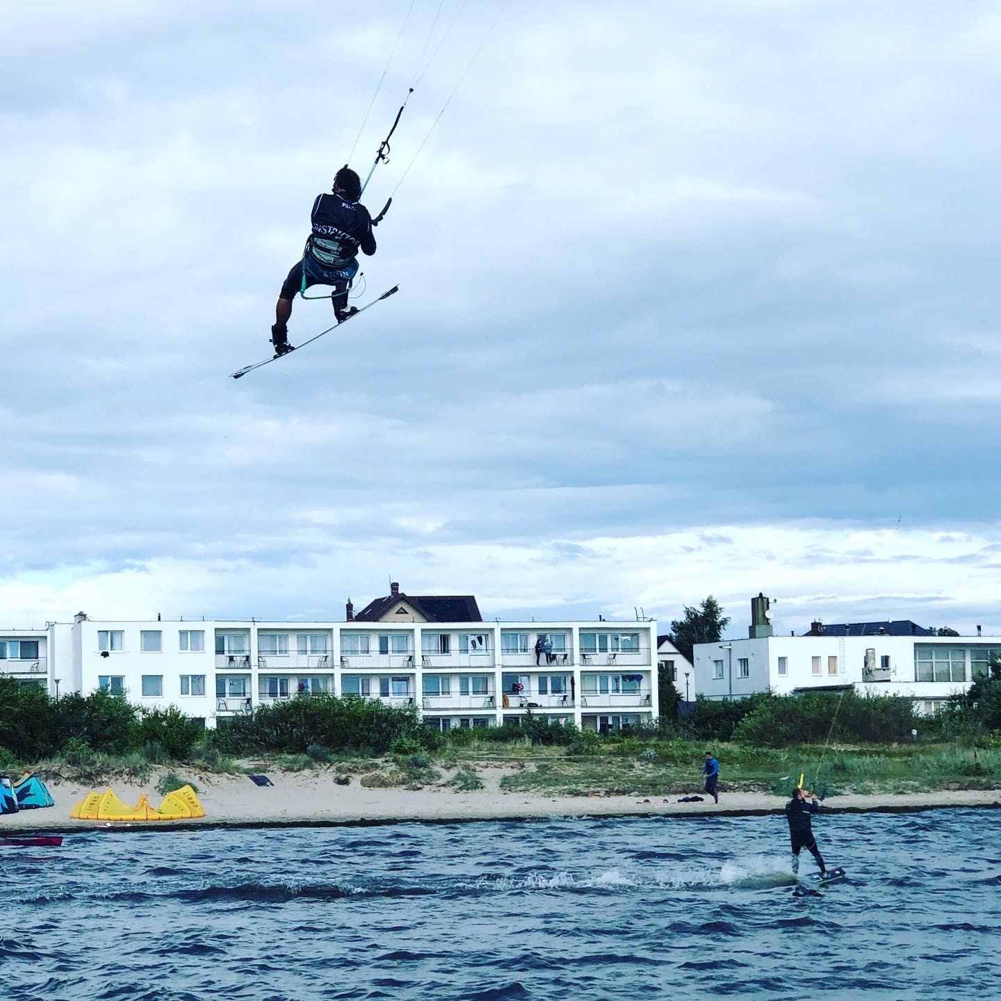Kitesurfer w locie nad wodą w Warszawie, w tle budynek mieszkalny i plaża.