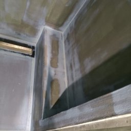 Profis - Narożnik pomieszczenia w trakcie prac wykończeniowych; ściany i sufit pokryte szarą warstwą z widocznymi śladami szpachlowania i wygładzania, widoczny fragment kratki wentylacyjnej.