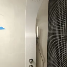 Biały panel prysznicowy z deszczownicą i słuchawką na tle ściany wyłożonej czarną mozaiką.