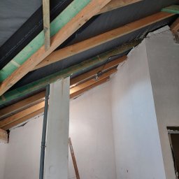 Profis - Wnętrze w trakcie remontu: surowy strop z drewnianych belek i czarnej membrany, ściany pokryte świeżo nałożoną, nierówną warstwą gładzi, wystająca rura kanalizacyjna, fragment drabiny i mężczyzna...