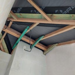 Profis - Sufit poddasza w trakcie wykończenia: widoczne drewniane belki konstrukcyjne, czarna membrana dachowa z nadrukiem, fragment instalacji elektrycznej i ściany po nałożeniu gładzi.