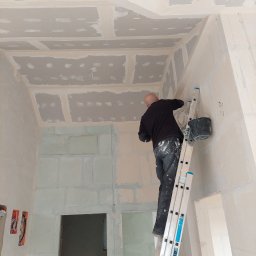 Profis - Pracownik na drabinie szpachluje narożnik ściany z płyt kartonowo-gipsowych, widoczny sufit z płytami i wiadro z masą szpachlową.